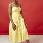 Anastacia-Renee stands, leaning back on one leg, smiling broadly. She wears a yellow dress and the wall is red behind her.