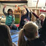 First grade students sit in a circle, holding their hands up