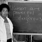 Audre Lorde stands next to a blackboard with these words written on it: "Women are powerful and dangerous."