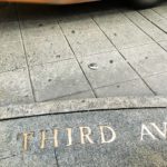 Photo of text embedded in the sidewalk in brass: "THIRD AVENIUE". A bus passes by just above, blurred out from the speed.