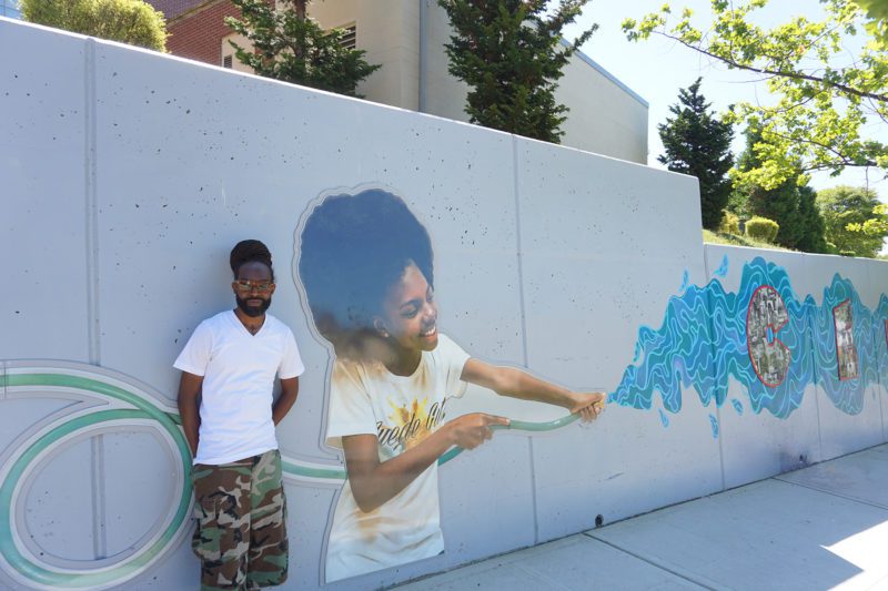 Freeman leans against the concrete retaining wall next to his piece: a young Black girl spraying a stream of water from a hose.