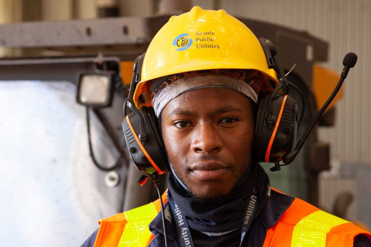 A Black man wearing a yellow hard hat, orange safety vest, and large headphones with a long mic angled up. Around his neck is a black gaitor.