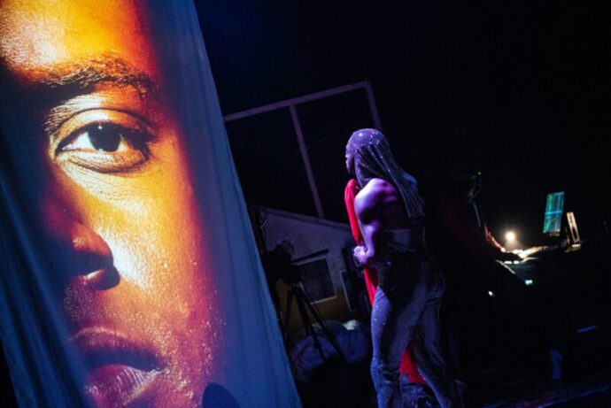 An actor on stage stands at a mic. In front of them is a projection of a Black person's face. From this angle, you can see the Stage Manager backstage at a computer terminal.