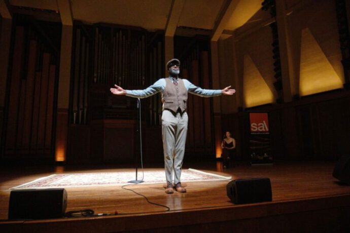 A Black storyteller stands with their arms spread wide on stage. Behind them is a mic stand on top of a square rug.