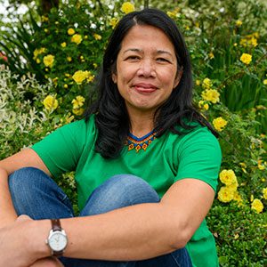 Carina A. del Rosario sits with her legs pulled up, arms around the knees. Behind her is a shrub with yellow flowers.