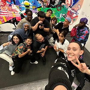 A group of youth and their adult staff member mug for a selfie in front of a mural wall.