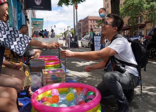 A street vendor hands a customer something outside of a storefront. Between them is a small inflatable pool filled with colorful orbs.
