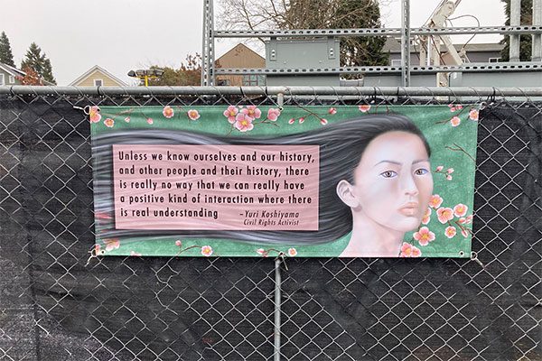 Artwork printed on a banner attached to a chain link fence. Illustration of an Asian woman with long black hair containing this quote: Unless we know ourselves and our history, and other people and their history, there is really no way that we can really have a positive kind of interaction where there is real understanding. Yuri Koshiyama, Civil Rights Activist