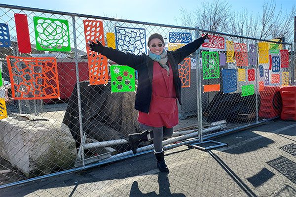 June stands on one leg, arms outstretched in front of her artwork. It's mounted on a chain link fence behind her: colorful vinyl panels with shapes and patterns cut out.