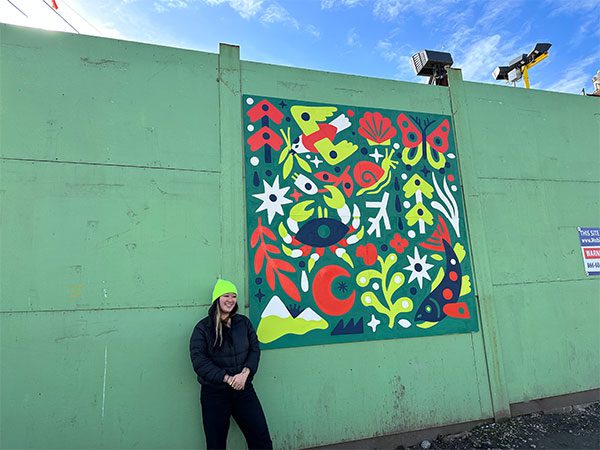Stevie stands next to her artwork that's mounted on a tall, wooden fence. Bright colors and shapes depict different animals, plants, and the natural environment with a limited color palette. 