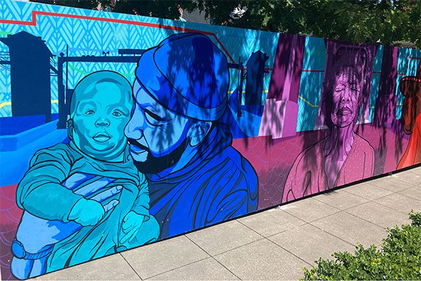 Brightly colored panels take up the entirety of the chain link fencing they're attached to. Three characters we can see are a man cuddling a baby, and an older woman with her eyes closed.