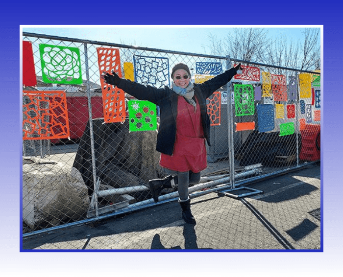 Woman standing with arms outstretched in front of a chain link fence with green, yellow, blue, red, and orange artwork. artwork are rectangles with shapes cut out.