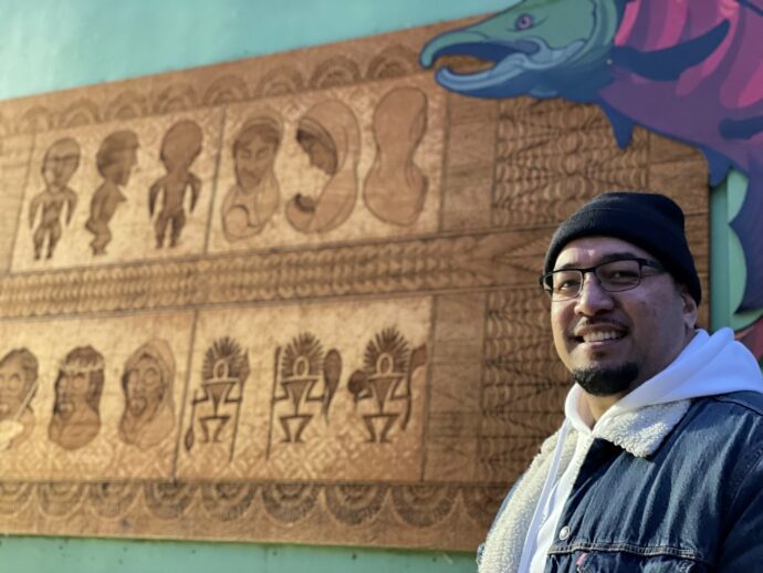 Artist Toka Valu smiles into the camera with his mural in the background. A salmon appears above him.