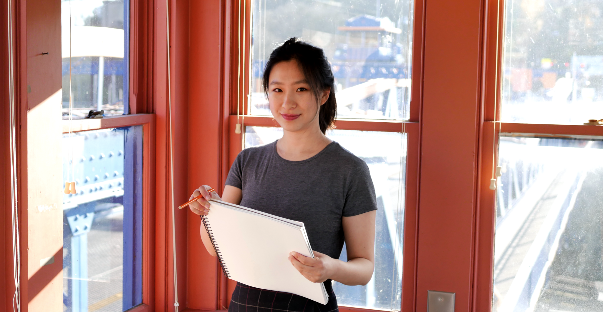 A person with brown eyes and brown hair pulled back in a ponytail wears a grey t-shirt and holds a drawing pad and pencil. There are burnt orange colored window frames around them and a blue bridge in the background behind them.