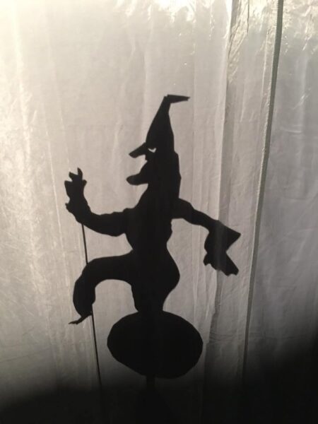A shadow puppet is shown in profile with a pointed hat, a long chin and nose, and balanced on a ball.