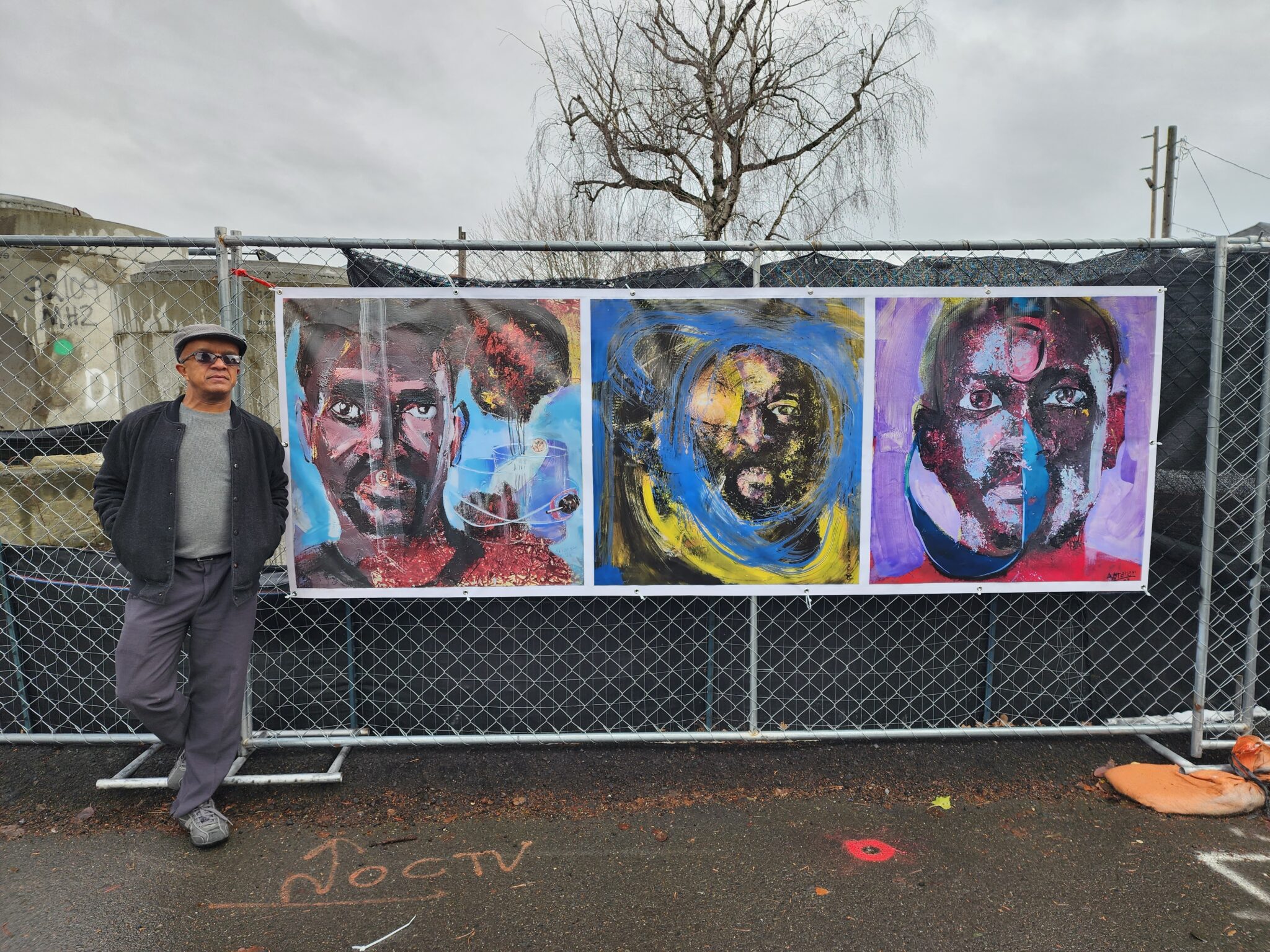 Man stands outside next to horizontal painting of three portraits hung on chain link fence