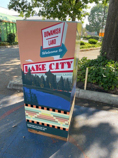 A vinyl wraparound artwork on a utility box depicts a retro road sign that says Duwamish Land, Welcome to Lake City. Below is a river, trees, colored patterns, and a dedication: "In honor of those that were here before me".