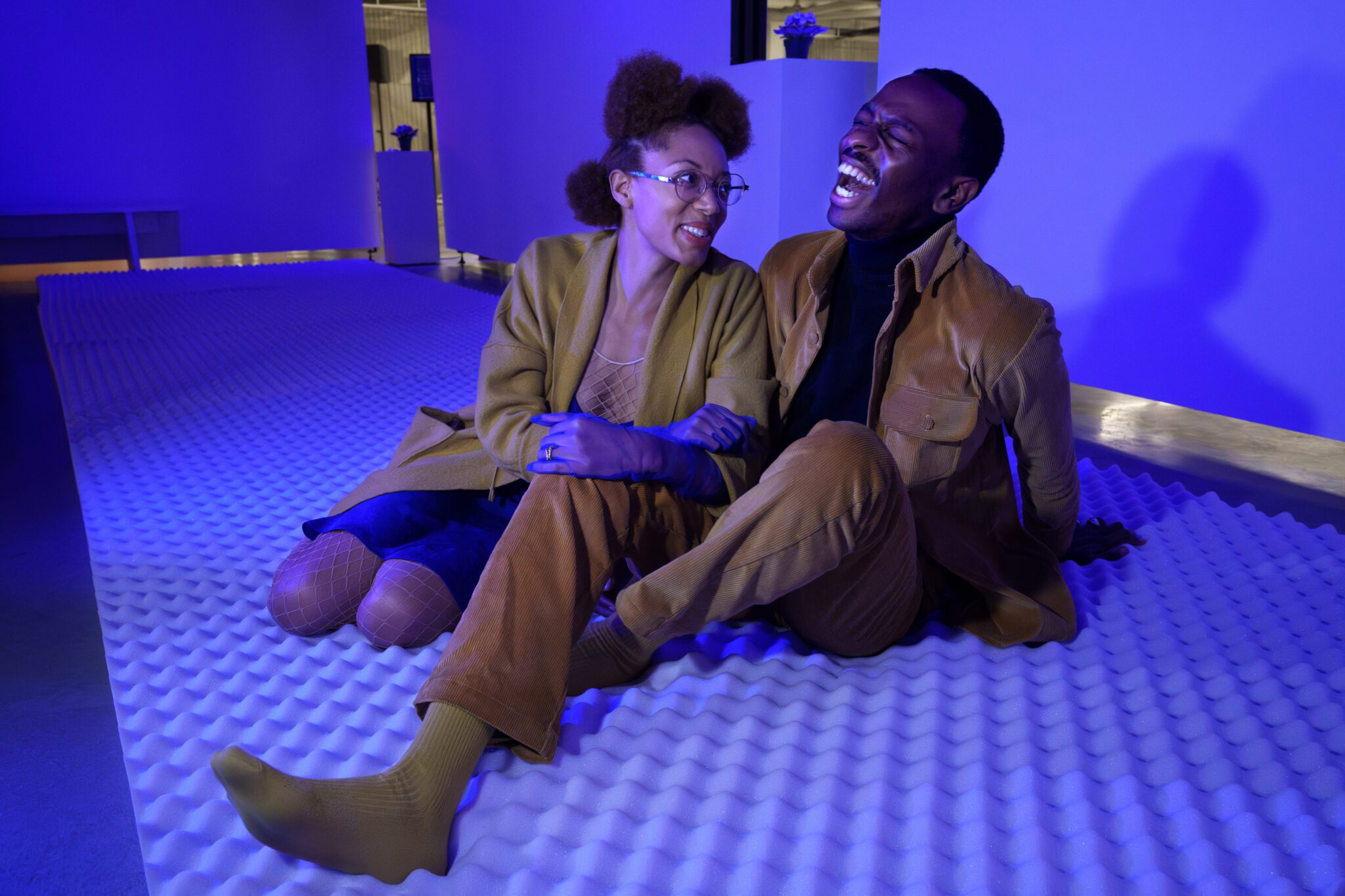 Black woman and Black man sitting together and laughing. Sitting on white foam and background is white walls.