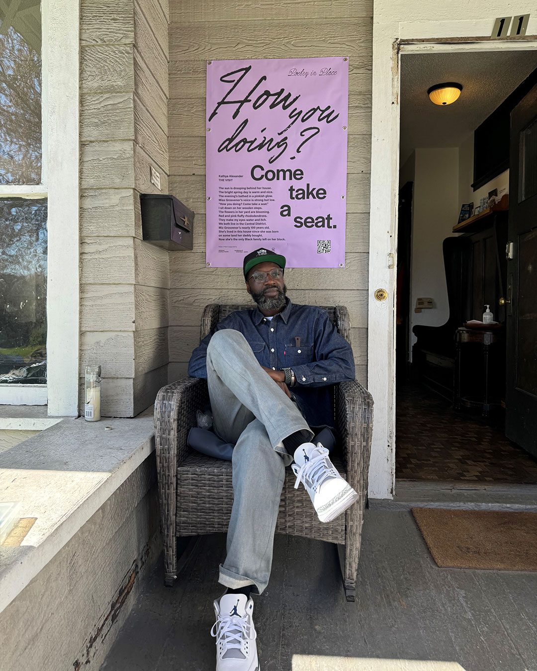 A Black man sits on the porch of Wa Na Wari below the poem by Kathya Alexander that's mounted on the wall.