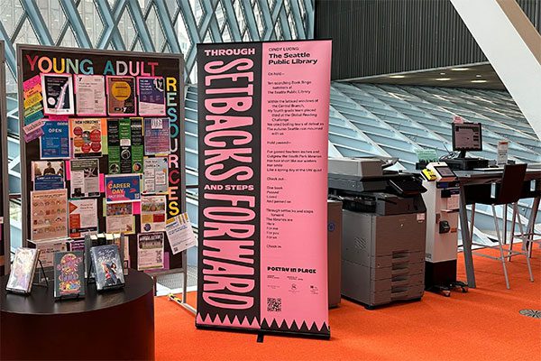 Black and pink standing banner of Cindy Luong's poem next to a library resource board.