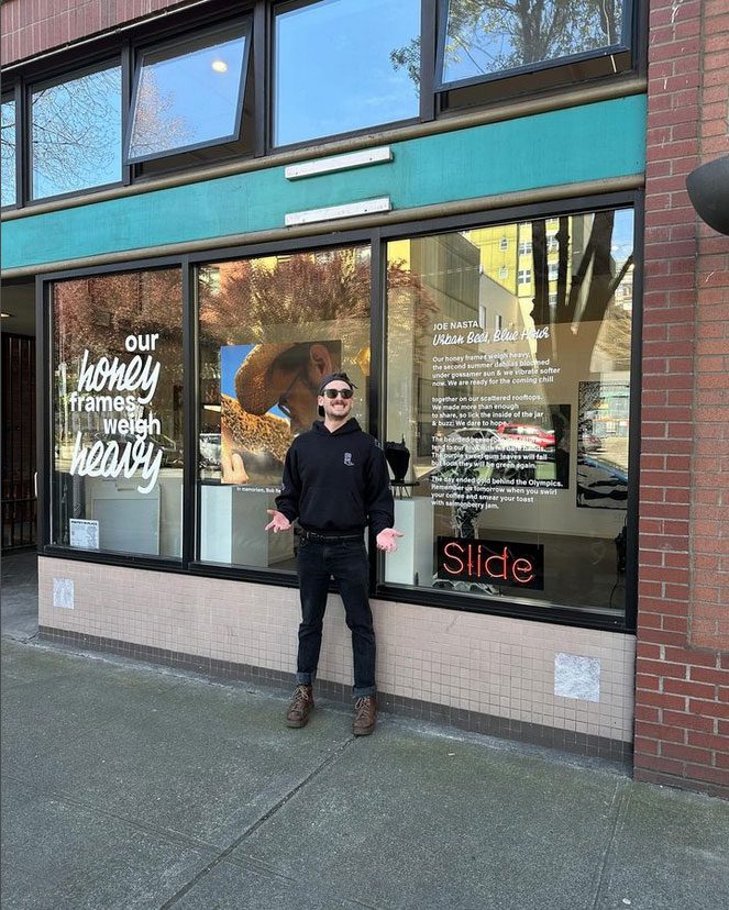 Joe stands smiling outside a gallery with hands out, palms upward. Behind on the window is zir poem in vinyl.