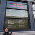 Photo of Bryna standing in front of building window that has vinyl installation of poem under friends of little Sài Gòn neon sign.