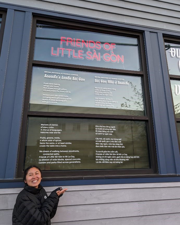 Photo of Bryna standing in front of building window that has vinyl installation of poem under friends of little Sài Gòn neon sign.
