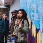 A Black woman with dreads talks to a group gathered around a bright mural that she painted.