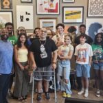 Group photo of Fresh perspectives cohort with artist Damon Brown with many graphics hung on wall behind them.