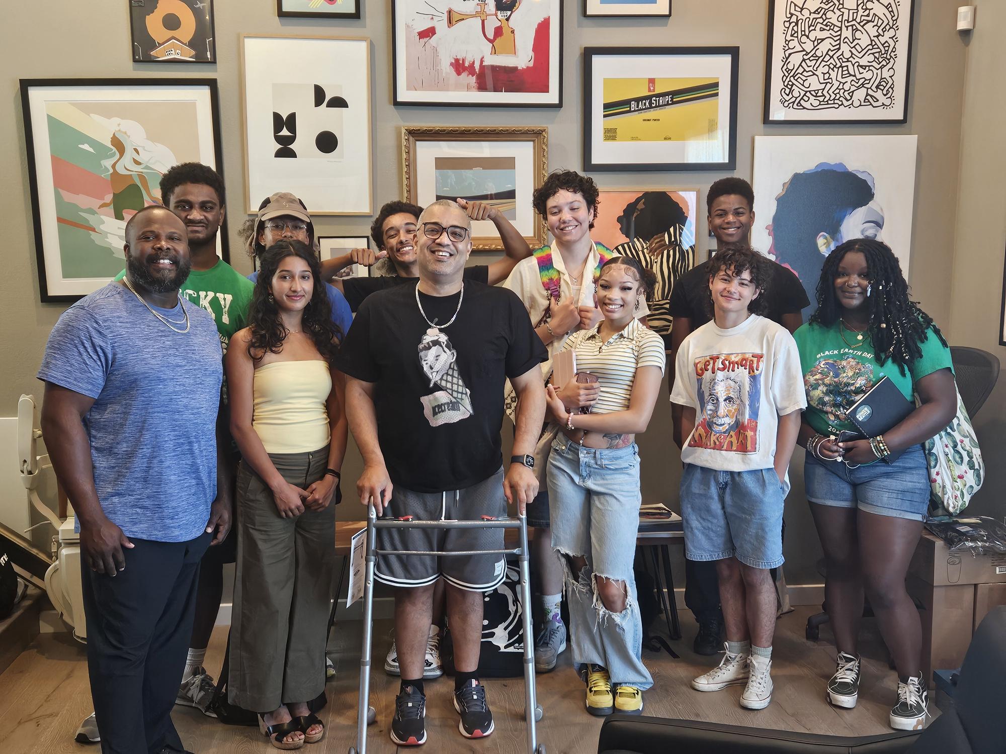 Group photo of Fresh perspectives cohort with artist Damon Brown with many graphics hung on wall behind them.