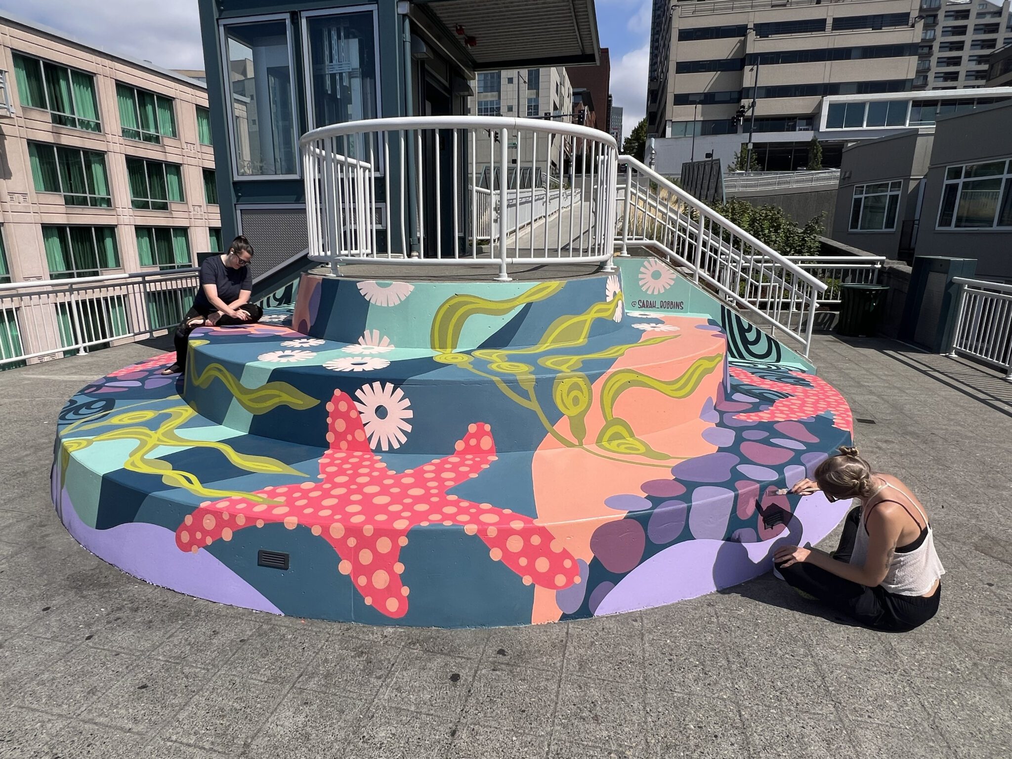 Two women paint colorful, underwater themed mural on large, rounded steps