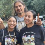 Two young Native dancers with plaited hair smile. A taller artist rests his hands on their shoulders and makes a peace sign.