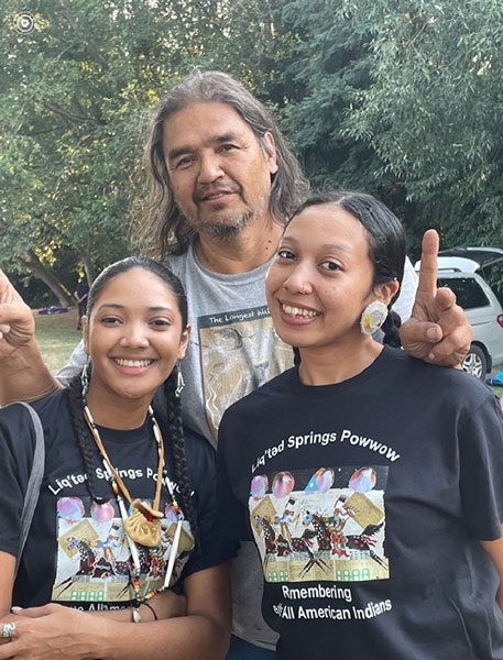 Two young Native dancers with plaited hair smile. A taller artist rests his hands on their shoulders and makes a peace sign.