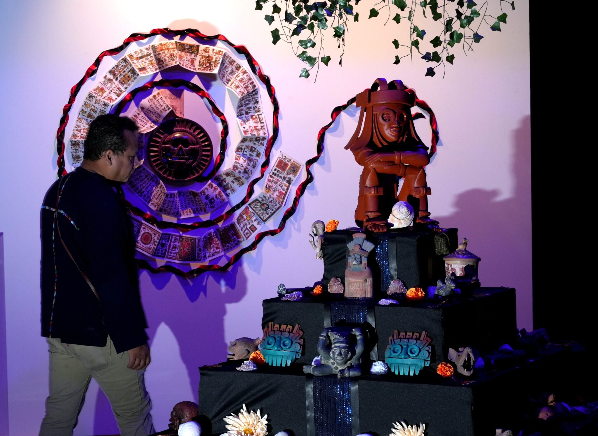 A man walks through gallery artwork of a pyramid holding many objects on it's steps and an unfolding spiral on the wall