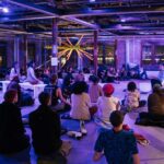 A large group of people sit on the floor and benches in the gallery, looking at a sound bath performance in the center of the space.