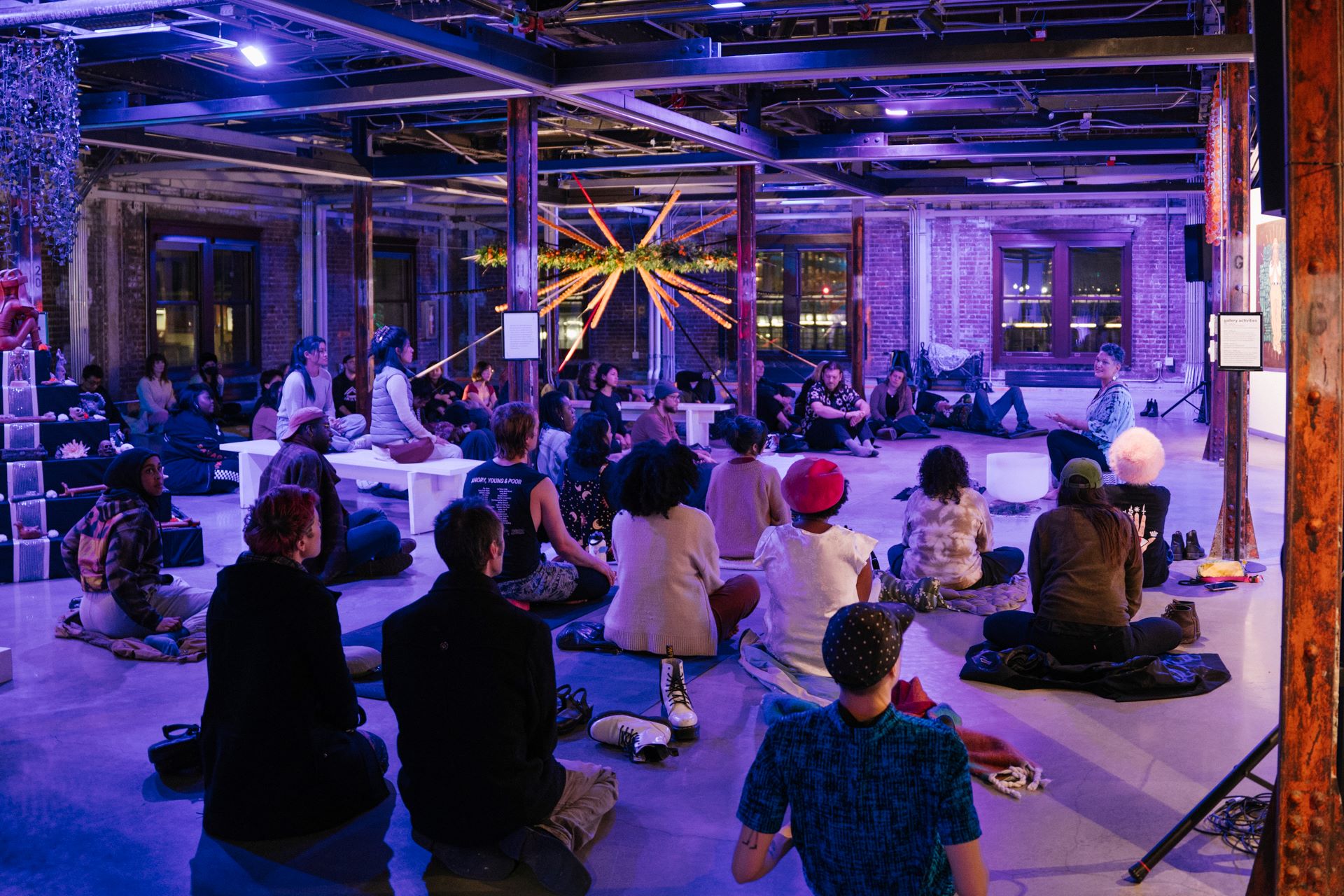 A large group of people sit on the floor and benches in the gallery, looking at a sound bath performance in the center of the space.