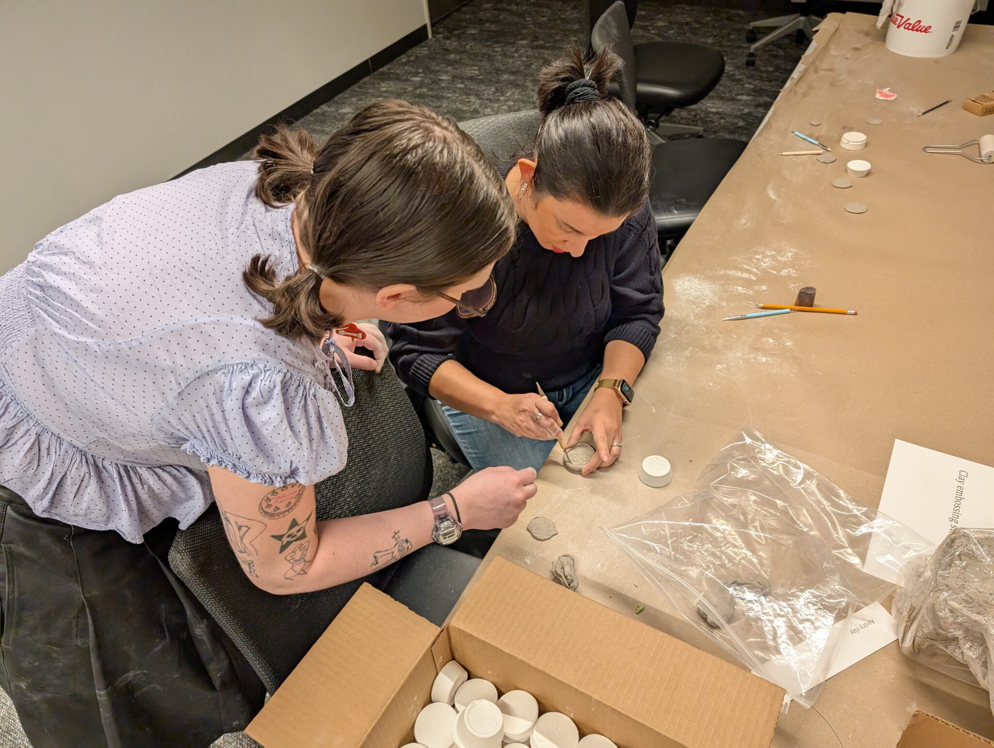 Two women carve a small clay object together at a table