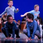 Young children dance on stage at Pacific Northwest Ballet. In the foreground, two of them crouch with their hands on the ground in front of them. They're looking at each other and smiling. Behind, two more run, and a third crouches.