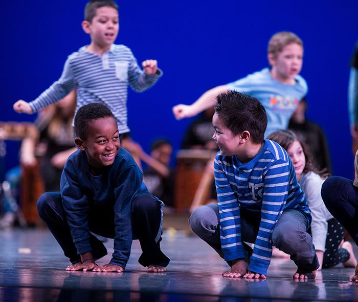 Young children dance on stage at Pacific Northwest Ballet. In the foreground, two of them crouch with their hands on the ground in front of them. They're looking at each other and smiling. Behind, two more run, and a third crouches.