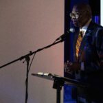 An elder Black man in a suit speaks into a mic in the gallery.