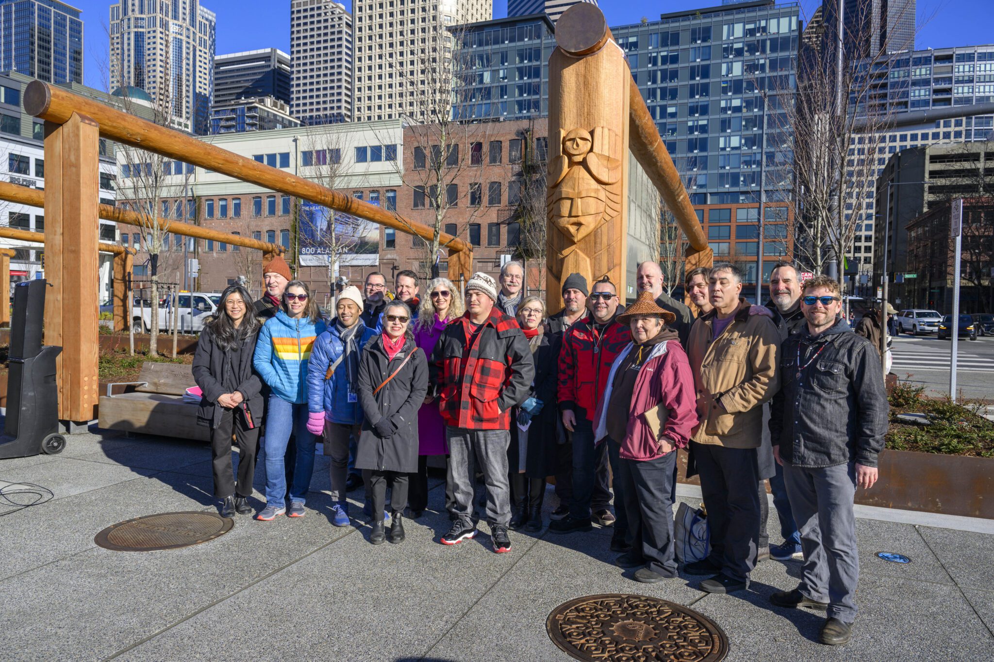 A group of artists and city officials stand in a group photo Infront of the wooden arches with tribal carvings.