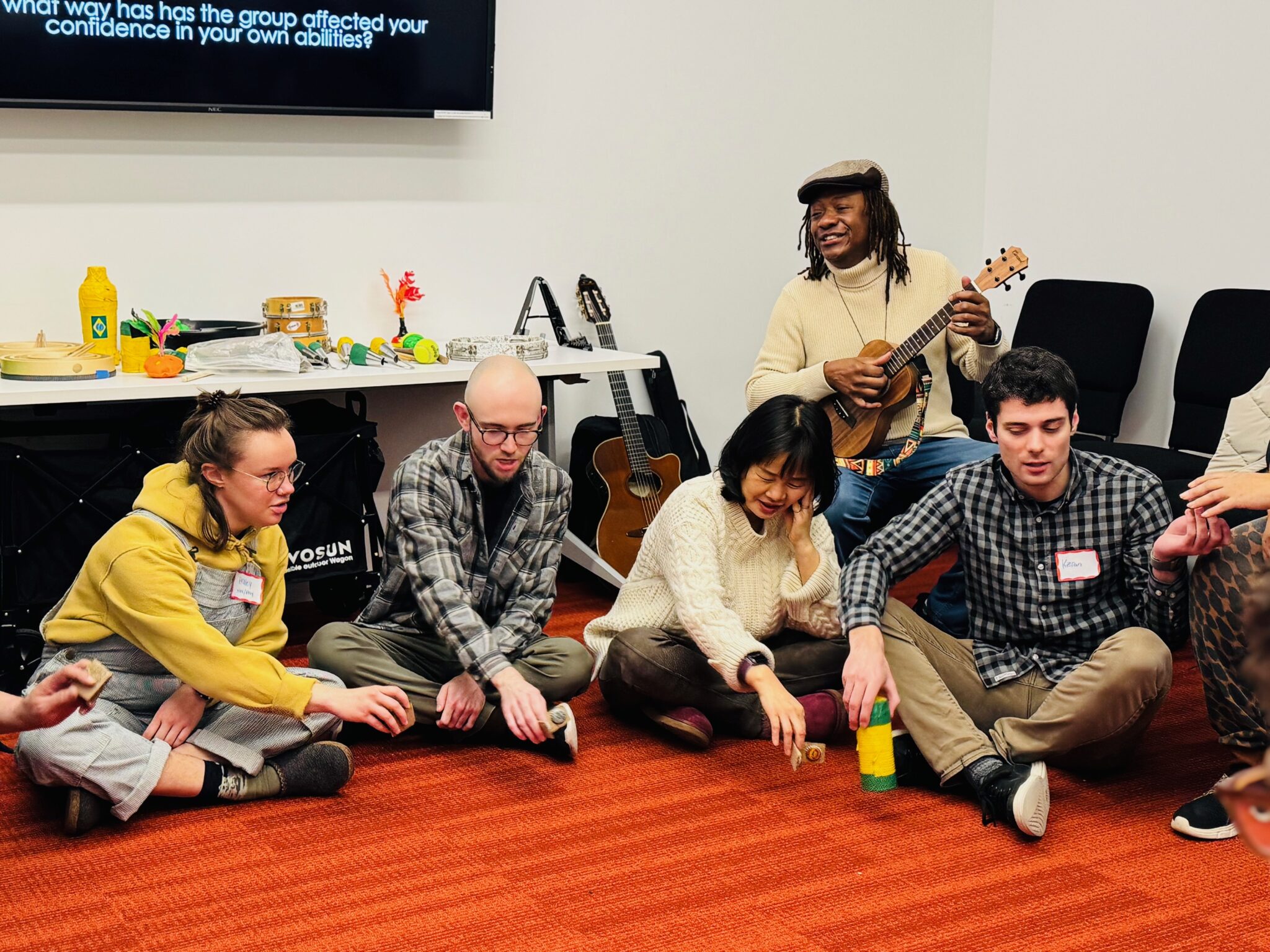 four teaching artists sit on the floor and have their hands in the middle while a man sits near them and stums a guitar with a soft smile.