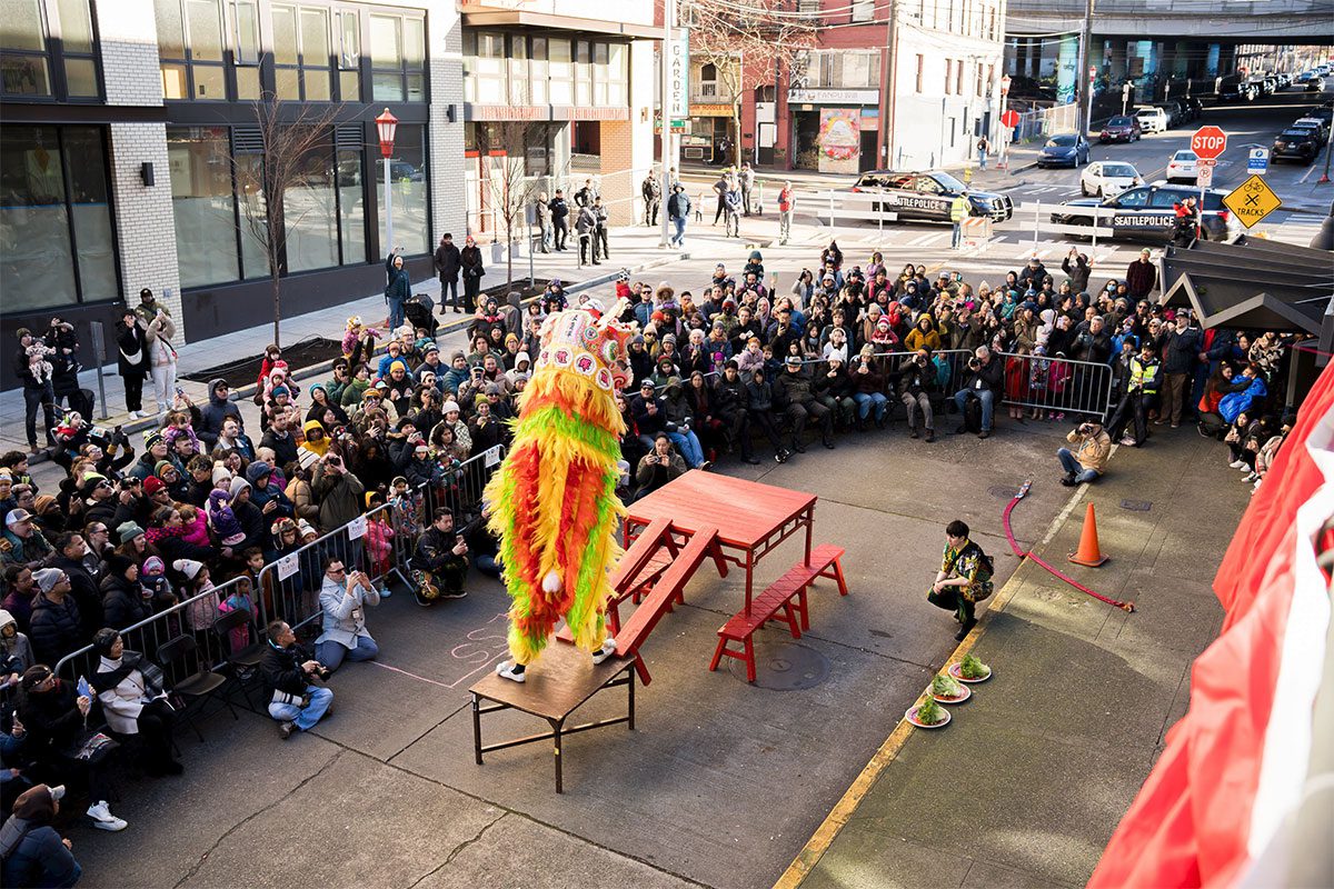 A lion dancer walks up an elevated platform. Crowds of people watch on the closed off street in the ID.