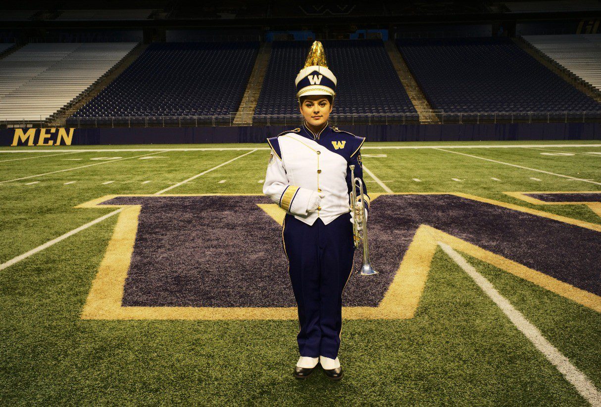 A young woman stands alone on the University of Washington football field dressed in a band uniform and holding a trumpet.