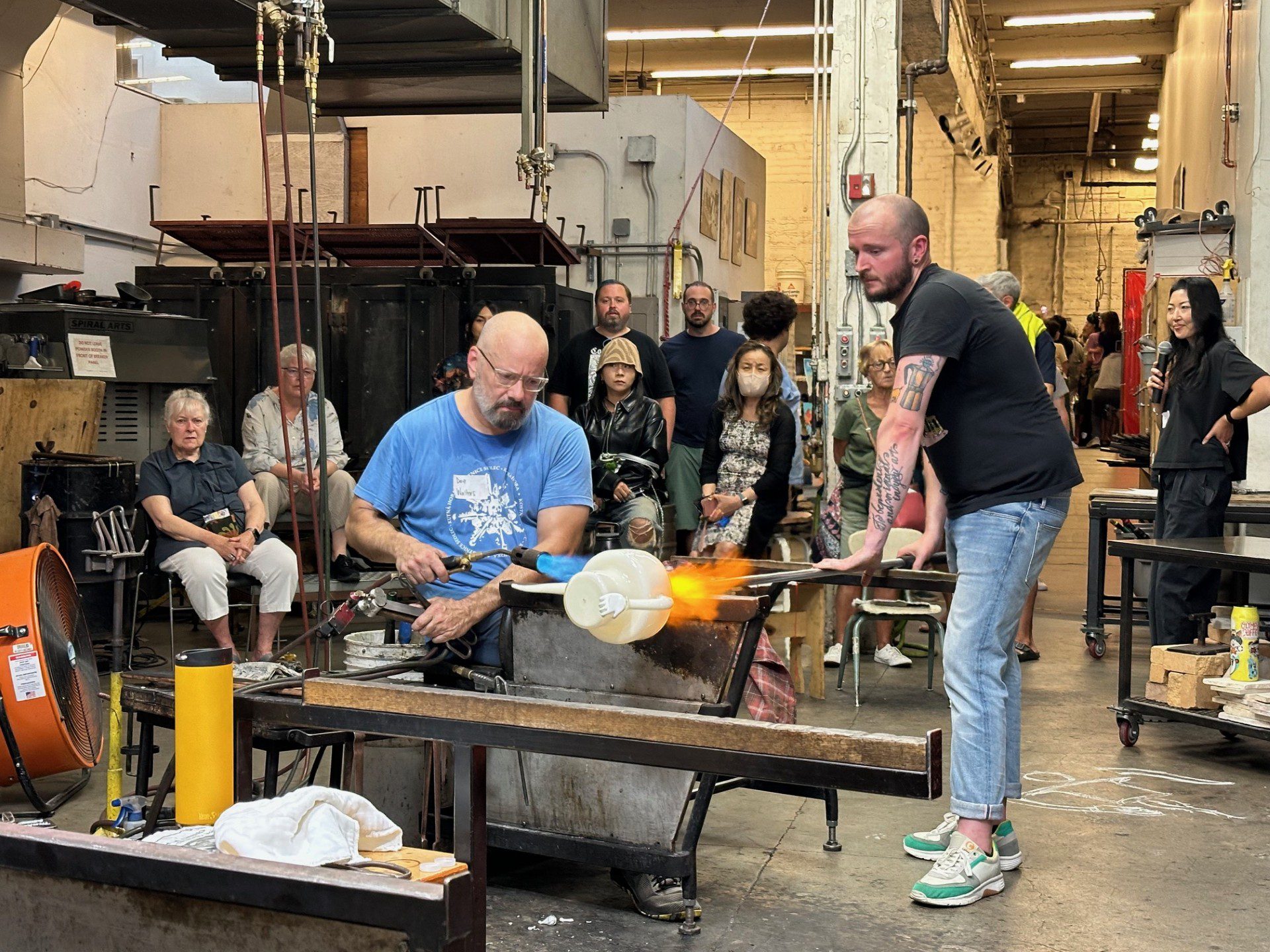 Two bald white men demonstrate glassblowing in the hot shop