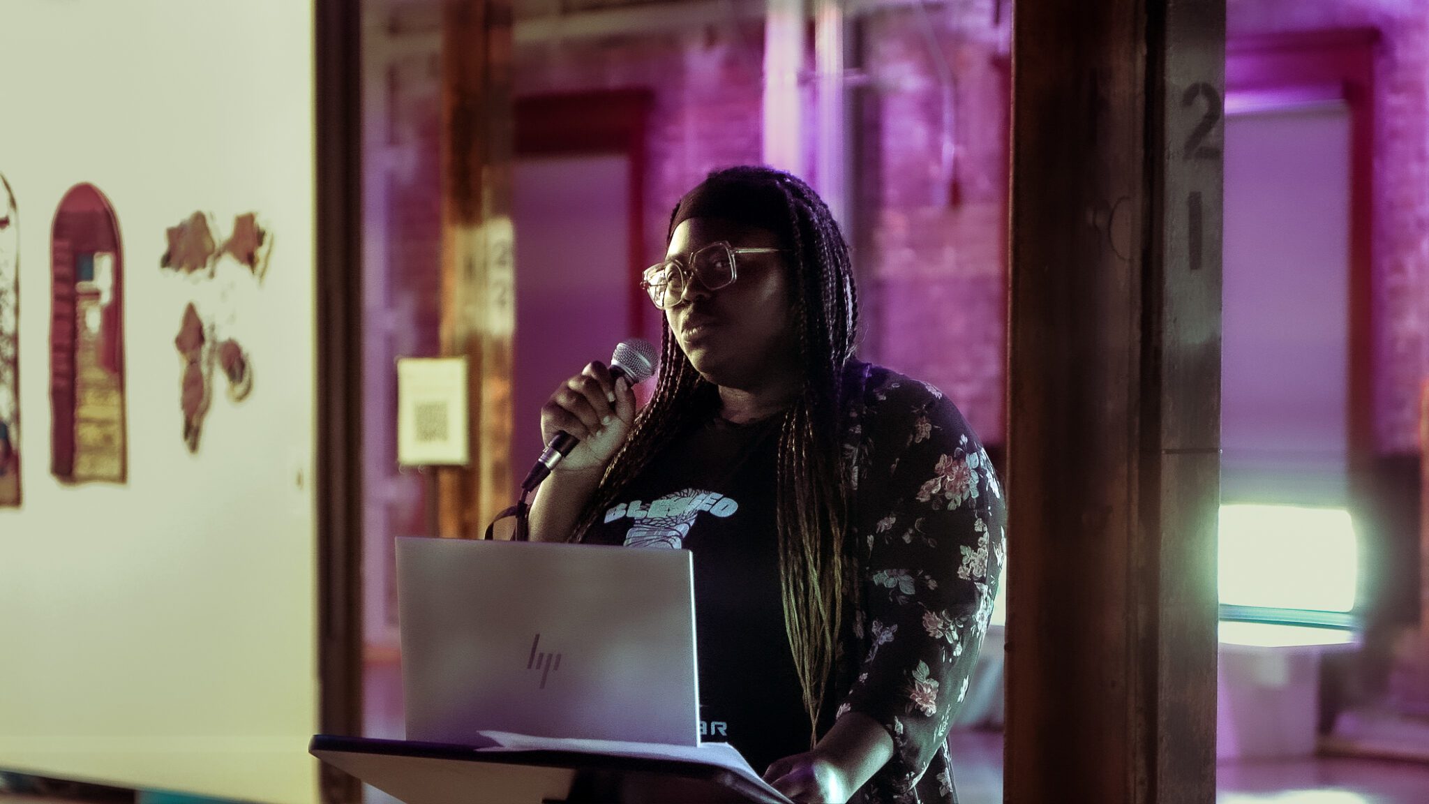 A teen with glasses and long braids speaks into a microphone while looking at the camera.