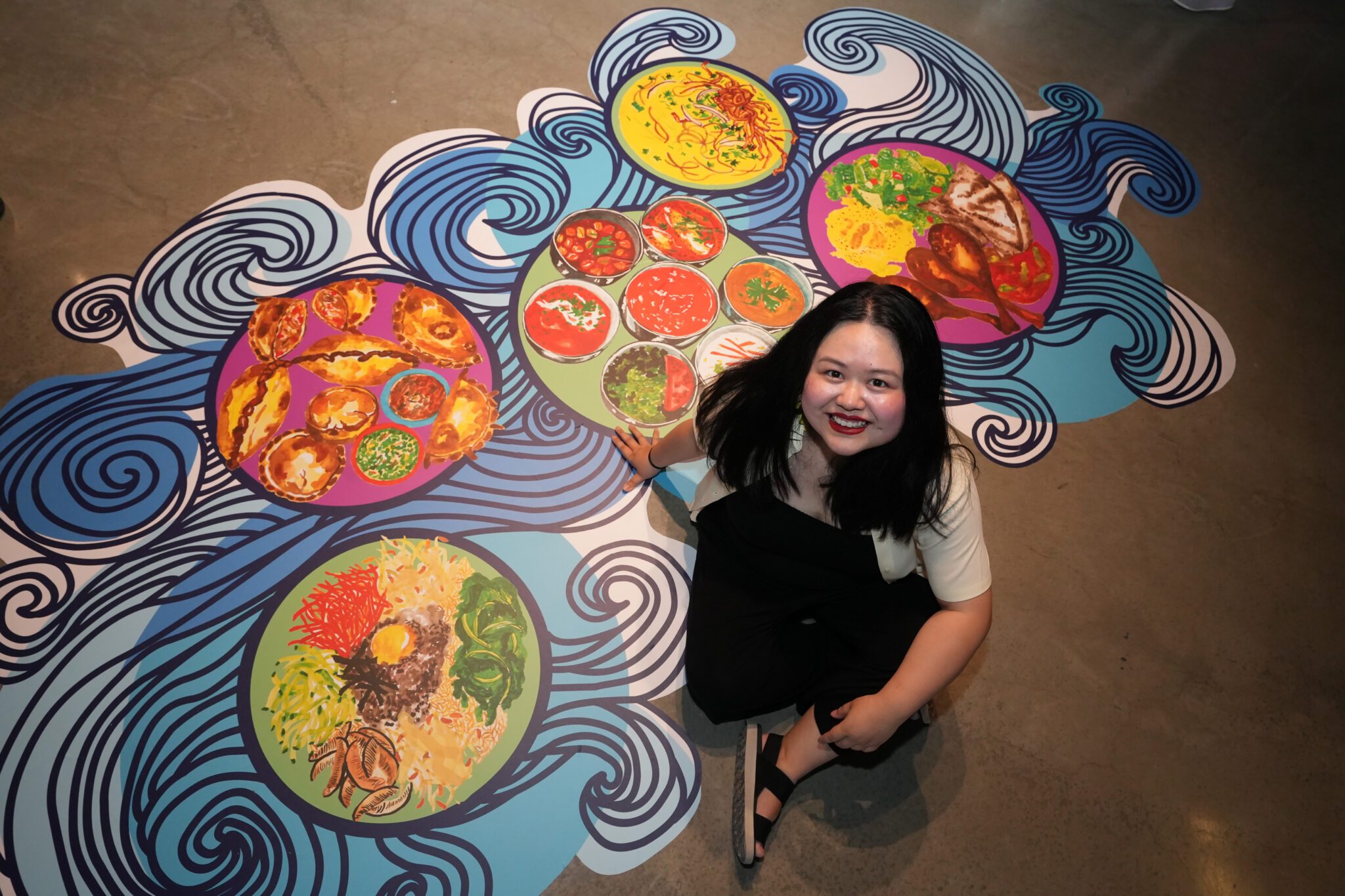 Artist Sophia Fang smiles on the ground next to their work, a colorful vinyl of food.