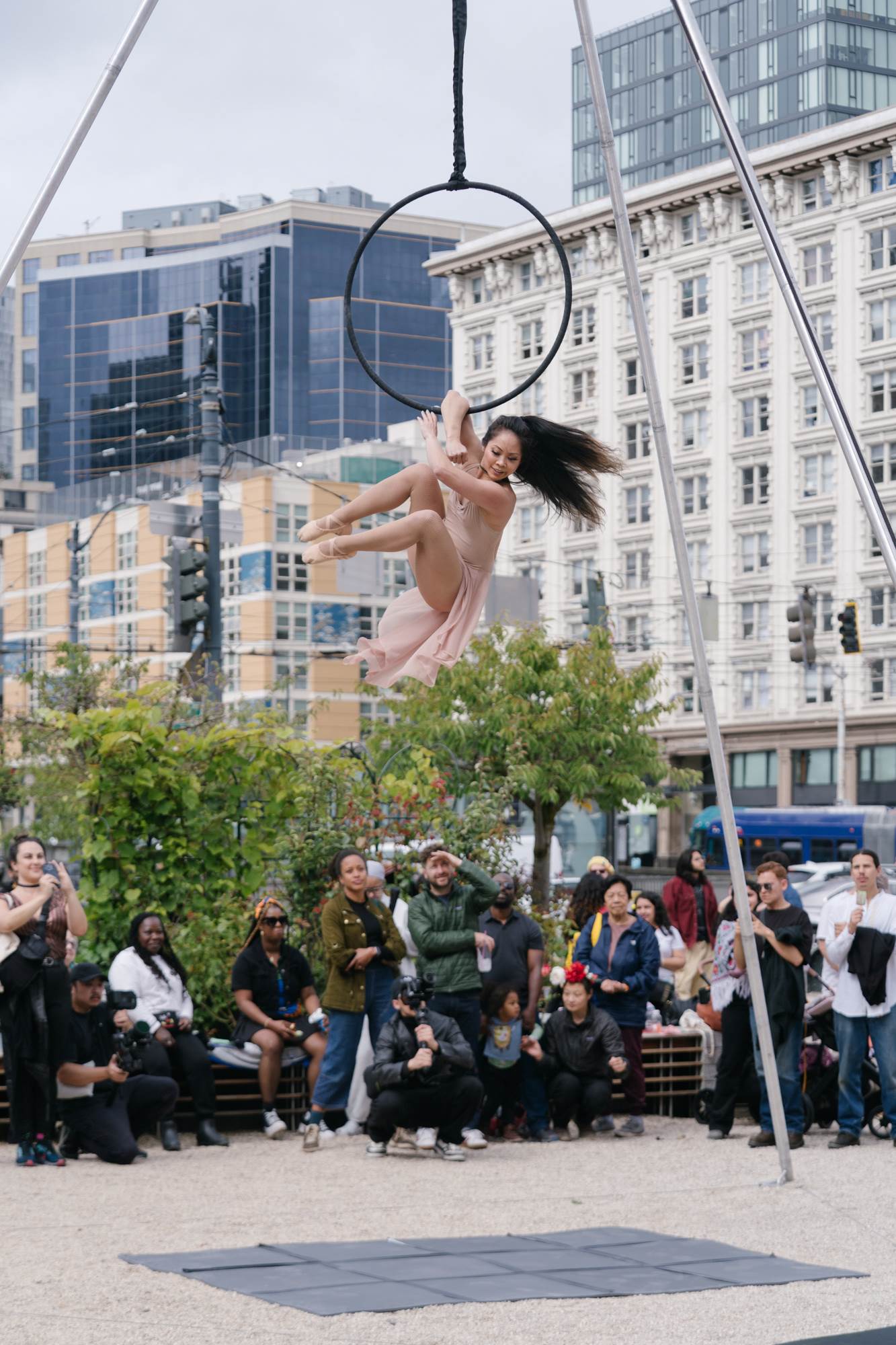 A femme performer in a pink dress and pointe shoes gracefully, hangs off an aerial hoop.