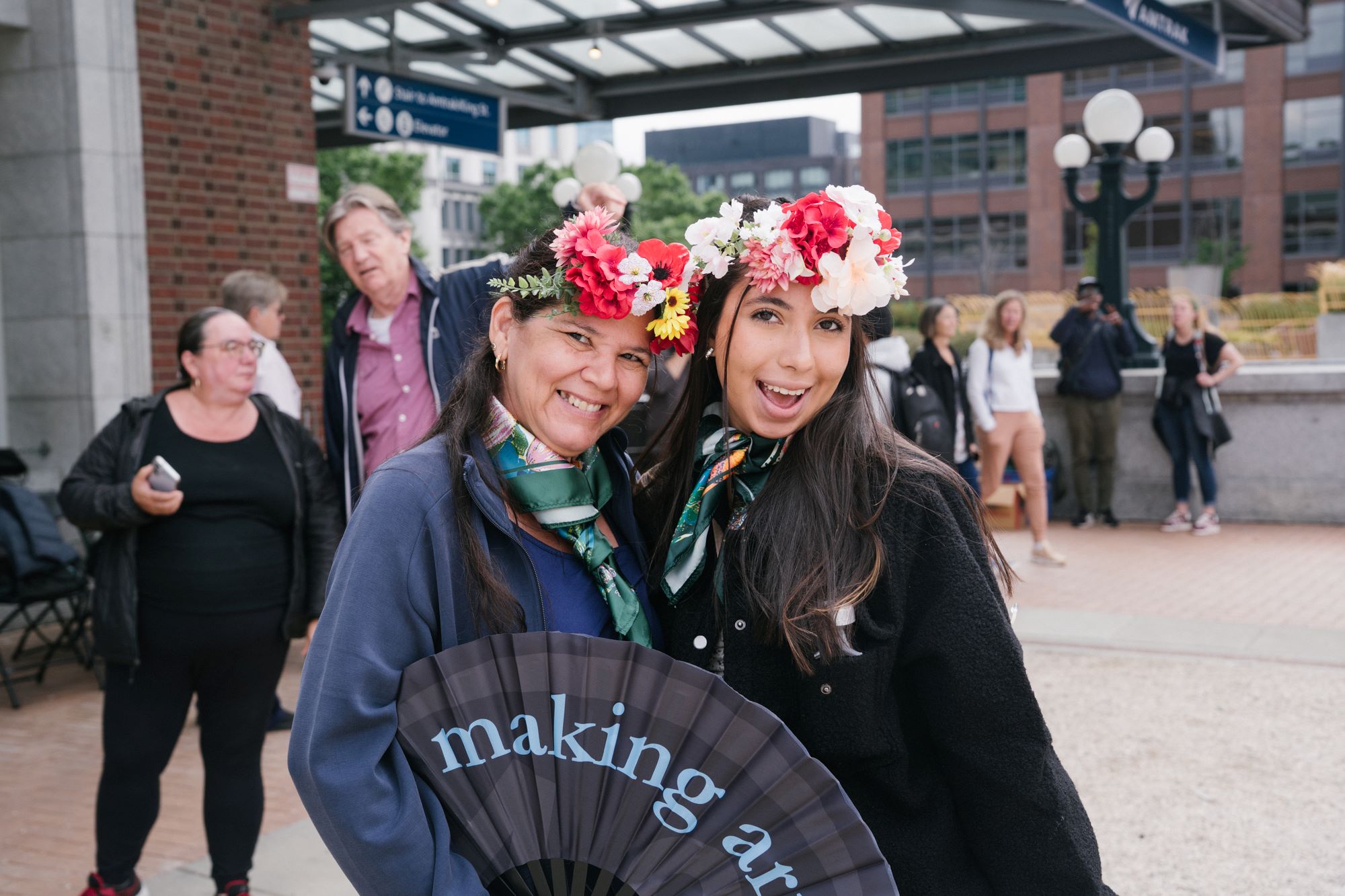 two women of different generations smile at the camera. They wear flower crowns and are holding a large fan that reads "making art"