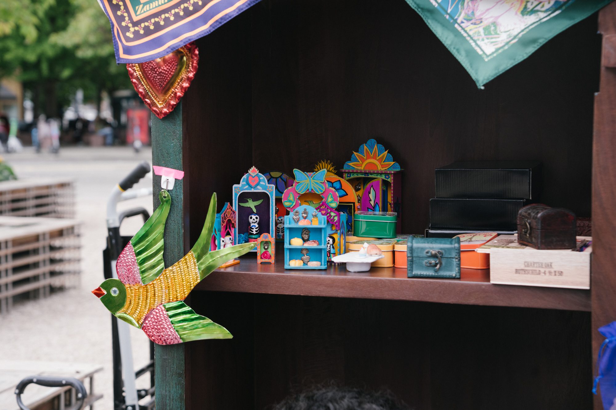A wooden shelf with a small colorful tin bird decoration hanging off it. On the shelf are a bunch of tiny handmade ofrendas and pieces of art.